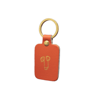 The Boob Key Ring - Coral is a rectangular orange leather keyring with a brass ring and stud, featuring a gold-embossed line drawing—an eye-catching accessory for your next Tallinn shopping trip or concept store discovery. Gate21.ee
