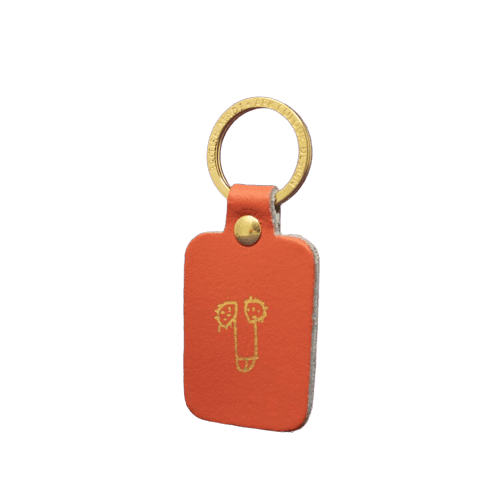 The Boob Key Ring - Coral is a rectangular orange leather keyring with a brass ring and stud, featuring a gold-embossed line drawing—an eye-catching accessory for your next Tallinn shopping trip or concept store discovery. Gate21.ee