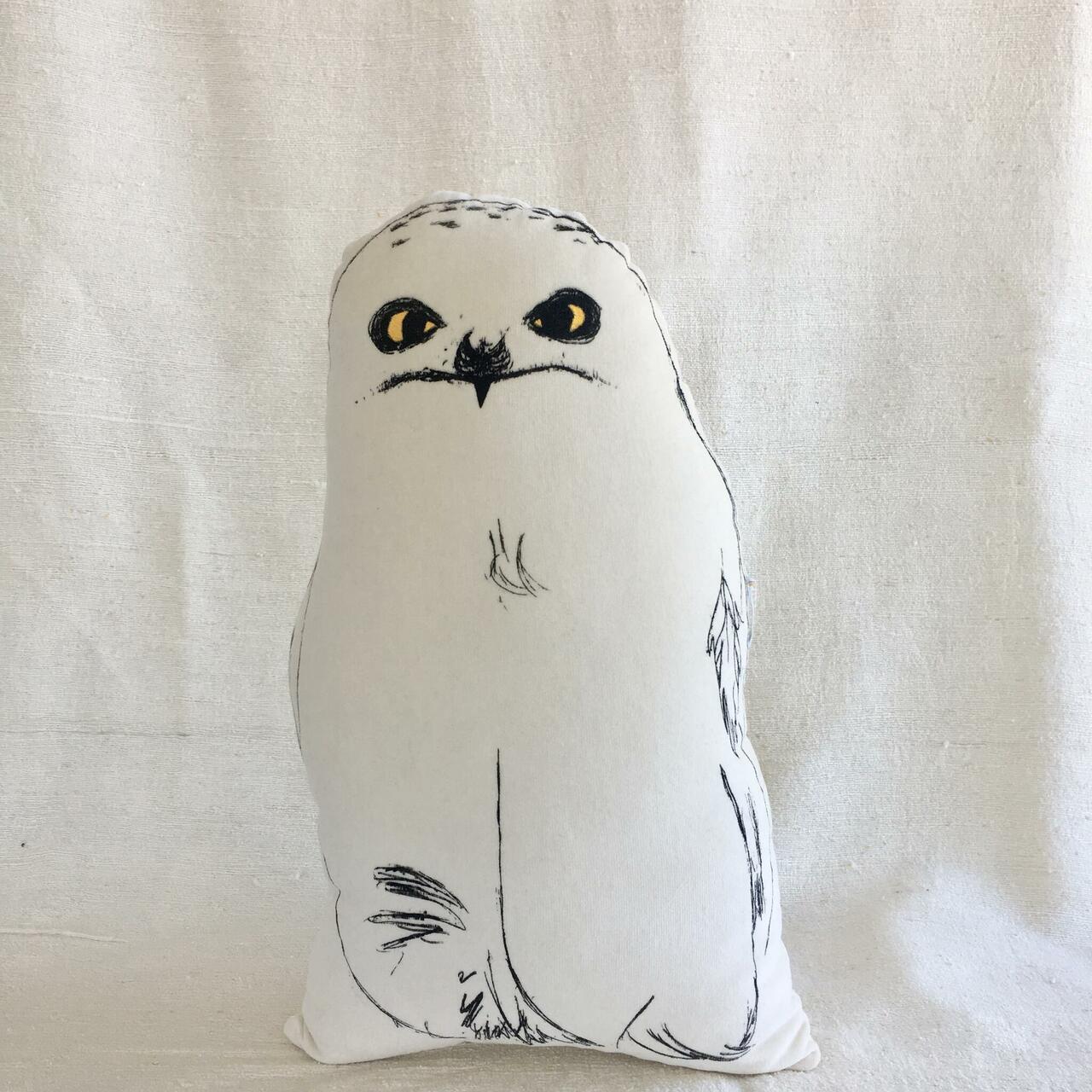 The Kakkud - Cushion - KÕIK is a white, owl-shaped pillow with minimalist black line art and yellow eyes, displayed upright against a light fabric background—an ideal choice for gift shops. Gate21.ee