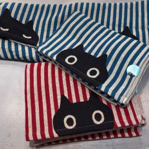 Three folded striped cloths with cartoon cat faces, curated for unique ideas, are stacked on a flat surface. Two blue and one red, each features a distinct cat expression and a small circular badge&mdash;perfect for any concept shop. Gate21.ee