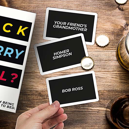 A hand holds an F'ck Marry Kill - Card Game card above a wooden table scattered with drinks, bottle tops, and other cards—great for sparking unique ideas on your next Tallinn shopping or gift shop adventure. Gate21.ee