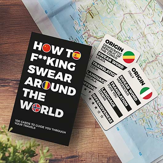 How to Swear around the World - Card Game is displayed on a table next to a map—ideal for your gift shop or concept store. Each card showcases international swear words with translations and flag icons in various languages. Gate21.ee