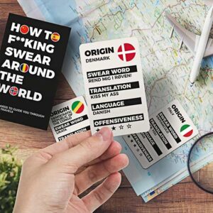 A hand holds a card from the "How to Swear around the World - Card Game" in front of a travel book—an ideal, unique gift for concept shops or shops specialising in playful, globally-themed items and Estonian design. Gate21.ee