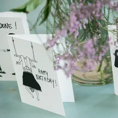 A Helen B Greeting Card - KÕIK, featuring Estonian design with a black and white drawing of a person hanging upside down from a swing and “Happy Birthday!” text—an ideal Tallinn shopping find. Gate21.ee