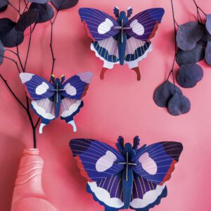 The Butterflies (Set of 3), featuring blue, purple, and white patterns, are displayed on a pink wall beside a curated dried leaf arrangement in a pink vase—an inspiring touch for unique home décor. Gate21.ee