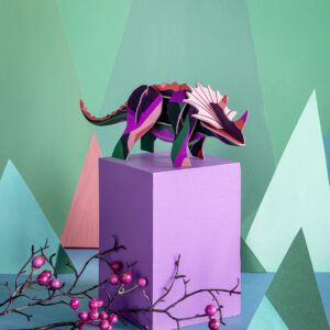 A vibrant paper Triceratops stands on a purple plinth, surrounded by pastel geometric shapes and pink-berried branches, capturing the playful vibe of an Estonian design concept shop against a green and blue backdrop. Gate21.ee