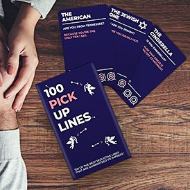 A hand rests on a wooden table beside the 100 Pick Up Lines - Trivia Card Game, available at a Tallinn concept shop of Estonian design. Three unique cards—The American, The Jewish One, and The Cinderella—are displayed. Gate21.ee