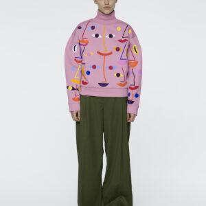 A person stands against a white background wearing the Pink - Pusa polo neck with an abstract face print, dark green wide-leg trousers, and black shoes—highlighting distinctive Estonian design for those craving unique fashion ideas. Gate21.ee