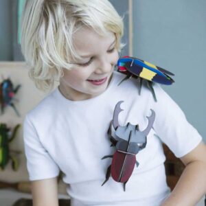 A child with light blond hair and a white shirt looks down, smiling at two large, colourful toy insects&mdash;one on their shoulder and one on their chest. Among them is the vibrant Stag Beetle from StudioROOF, adding a whimsical touch to the scene. The background is softly blurred with more toy insects visible. Gate21.ee