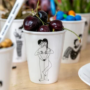 The Faces Cup is a white cup with dark cherries, featuring a black line drawing of two people—one standing behind and holding the other's chest. Ideal for Tallinn shopping or as a unique concept shop find, with blurred decorated cups in the background. Gate21.ee
