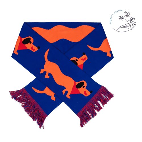 The Dachshund - Organic is a blue scarf with red tasselled ends, an orange dachshund dog pattern, and a small organic cotton label featuring a flower illustration in the top right corner, inspired by Estonian design. Gate21.ee