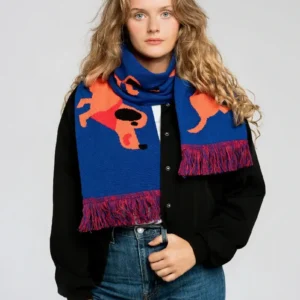 A woman with long wavy hair wears the Dachshund - Organic scarf, featuring orange dog patterns on blue, paired with a black button-up shirt and blue jeans, standing against a plain light background. Gate21.ee