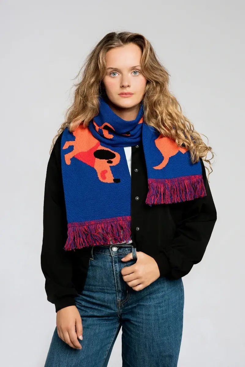 A woman with long wavy hair wears the Dachshund - Organic scarf, featuring orange dog patterns on blue, paired with a black button-up shirt and blue jeans, standing against a plain light background. Gate21.ee