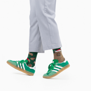 A person steps forward against a white background, wearing light grey trousers, green and white Adidas trainers, and mismatched Merrybara socks—one featuring a bear pattern and the other with a holiday design. Gate21.ee