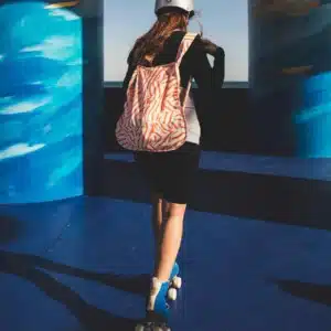 Wearing a helmet, zebra-striped rucksack, and roller skates, someone glides on a blue surface between pillars—sunlit shadows evoking the playful vibes of Peach Twist’s curated collection. Gate21.ee