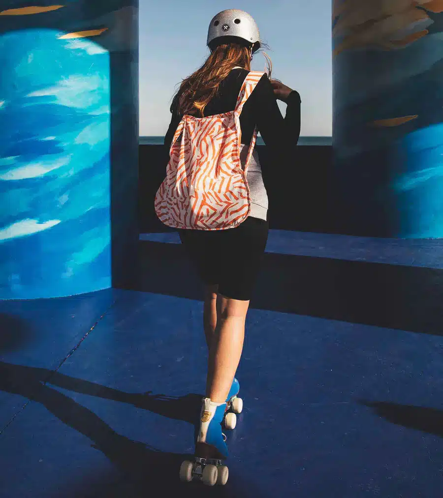 Wearing a helmet, zebra-striped rucksack, and roller skates, someone glides on a blue surface between pillars—sunlit shadows evoking the playful vibes of Peach Twist’s curated collection. Gate21.ee
