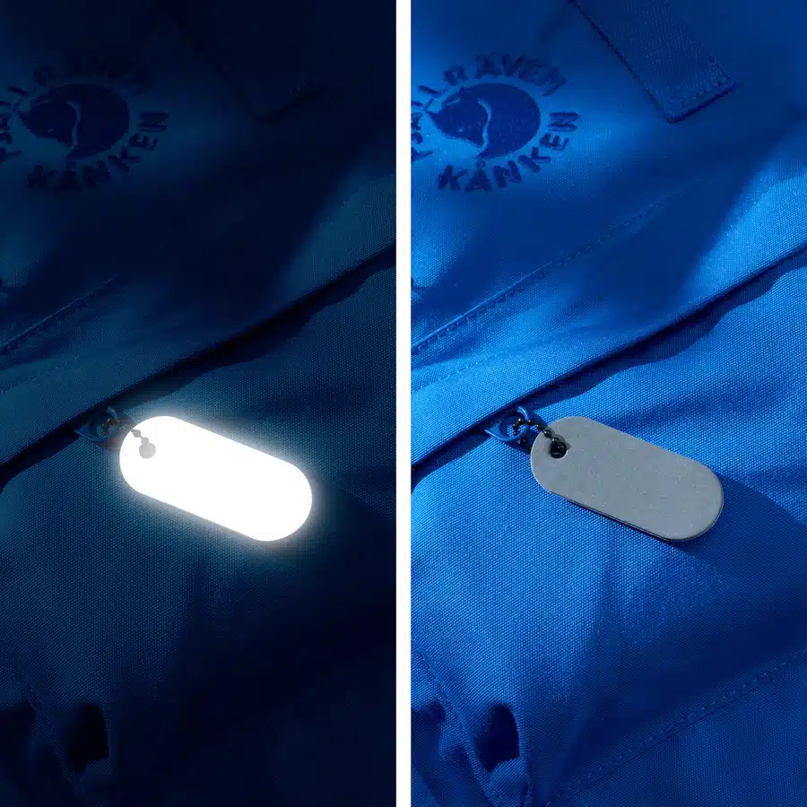 A comparison of a blue Fjällräven Kånken bag displays the Reflective Tag - Metallic Silver, inspired by Estonian design, attached to the zip—glowing on the left and appearing matte grey on the right. Gate21.ee