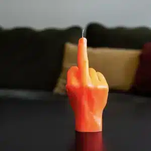 The F*ck You Candle - Neon Orange, shaped like a hand with the middle finger raised, stands boldly on a dark surface—a striking piece of Estonian design, ideal for quirky gift shops or your next Tallinn shopping trip. Gate21.ee