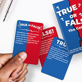 A hand displays cards from the True or False Trivia - Card Game, featuring white questions and answers on red and blue backgrounds. Part of the game’s box and a pen are also visible—ideal for discovering unique ideas at a concept shop. Gate21.ee