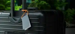 A black hard-shell suitcase with an extended handle and a Neon Orange Luggage Tag is pictured outdoors against blurred green foliage&mdash;an eye-catching Estonian design perfect for adding flair to your suitcase on Tallinn shopping trips. Gate21.ee