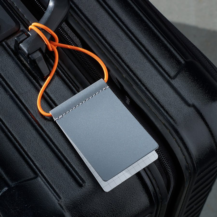A close-up of a black hard-shell suitcase with a Luggage Tag - Neon Orange attached. The tag features a neon orange loop, perfect for curated travel or Tallinn shopping trips, and has a white section with a grid pattern at the bottom. Gate21.ee