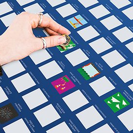 A hand with rings scratches off a square on the "100 Things to do with Dad - Bucket List" poster, which features curated illustrations in a grid, revealing colourful images of fun activities to share with Dad. Gate21.ee