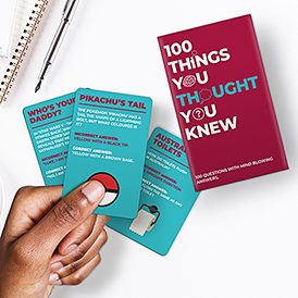 A hand holds three curated trivia cards beside the red 100 Things You Thought You Knew Trivia - Card Game box. With questions on Pikachu's tail, paternity, and Aussie toilets, it's perfect for gift shops or fun Tallinn finds. Notebook and pen included. Gate21.ee