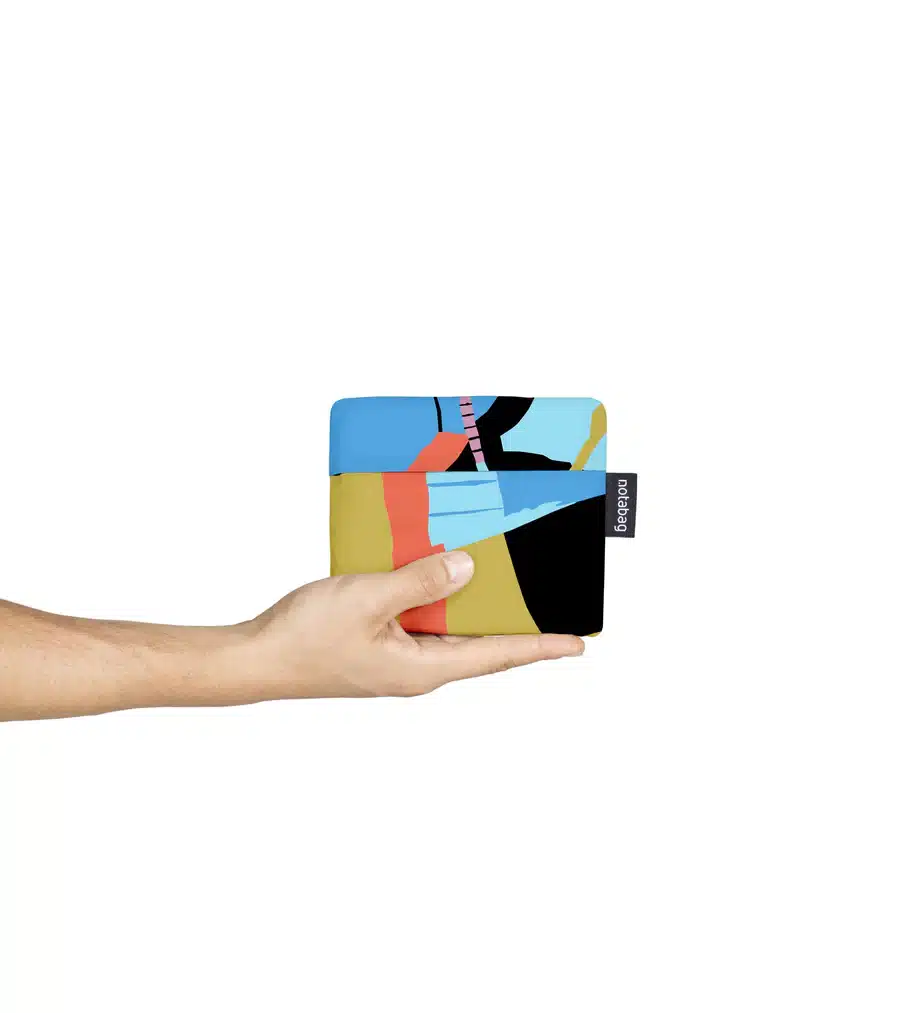 A hand holds the Roads - Notabag, a rectangular wallet with blue, red, black, and yellow abstract shapes plus a small black tag. Shot on a white background&mdash;perfect for curated gift shops seeking unique designs. Gate21.ee