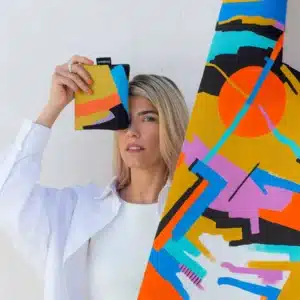 A blonde woman holds up a colourful Roads - Notabag in front of her eye, standing next to vibrant patterned fabric—showcasing the creative and modern style you’d find in a curated gift shop. Gate21.ee