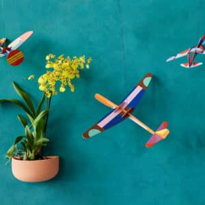 A yellow flowering plant in a wall-mounted pot stands out against a teal wall, complemented by three Giant Glider Plane models hanging nearby—an ideal way to showcase curated Estonian design with your Tallinn shopping finds. Gate21.ee