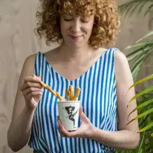 A woman with curly red hair in a blue and white striped sleeveless top holds the Girl Power Cup filled with breadsticks, looking down as she picks one up. A potted plant is partially visible on the right. Gate21.ee
