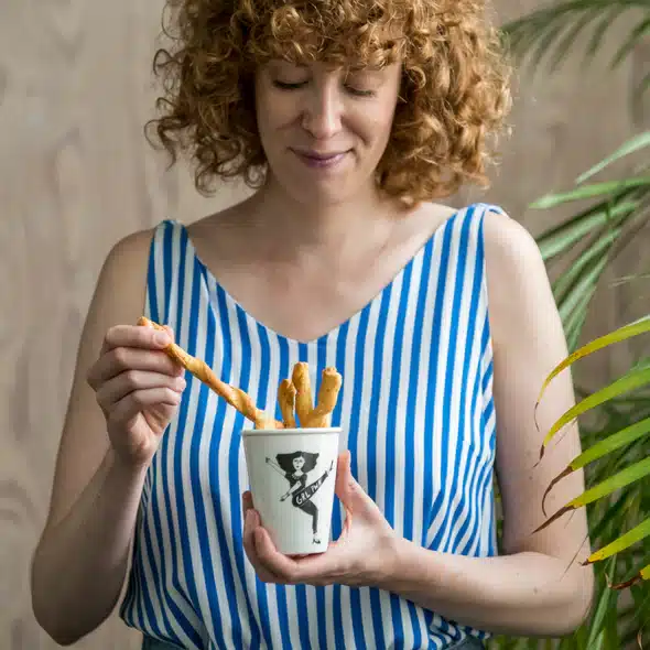 A woman with curly red hair in a blue and white striped sleeveless top holds the Girl Power Cup filled with breadsticks, looking down as she picks one up. A potted plant is partially visible on the right. Gate21.ee