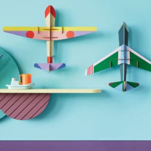 Two jet aeroplane models in vibrant colours are displayed on a light blue wall next to a shelf with a small jug, cup, orange flowers, and a mirror—unique inspiration for any concept shop or Tallinn shopping experience. Gate21.ee