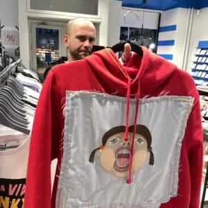 The Riivo Pusad red hoodie, with its large stitched cartoon face patch, stands out as a curated piece of Estonian design, catching the attention of a sceptical onlooker in the background. Gate21.ee