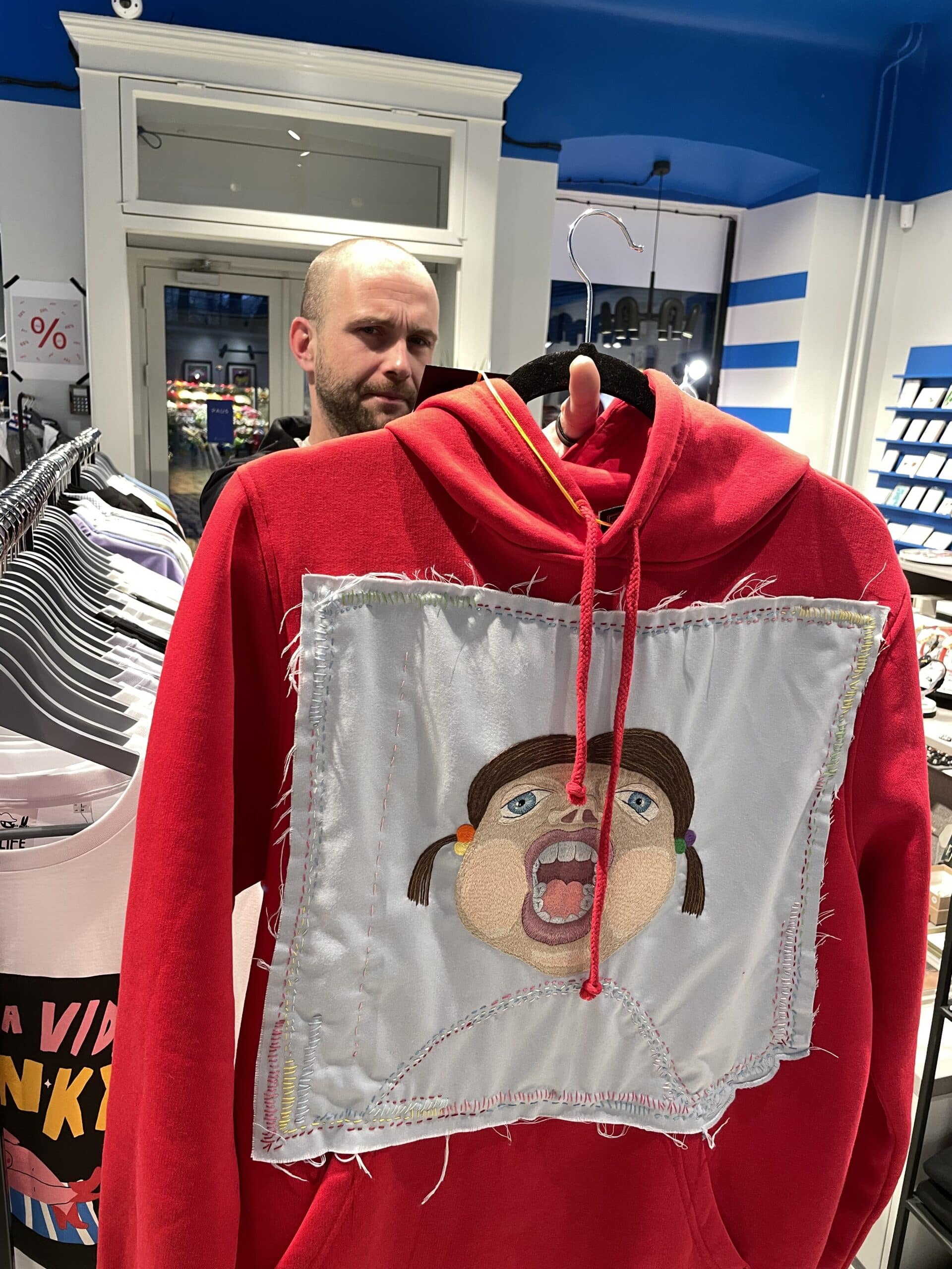 The Riivo Pusad red hoodie, with its large stitched cartoon face patch, stands out as a curated piece of Estonian design, catching the attention of a sceptical onlooker in the background. Gate21.ee