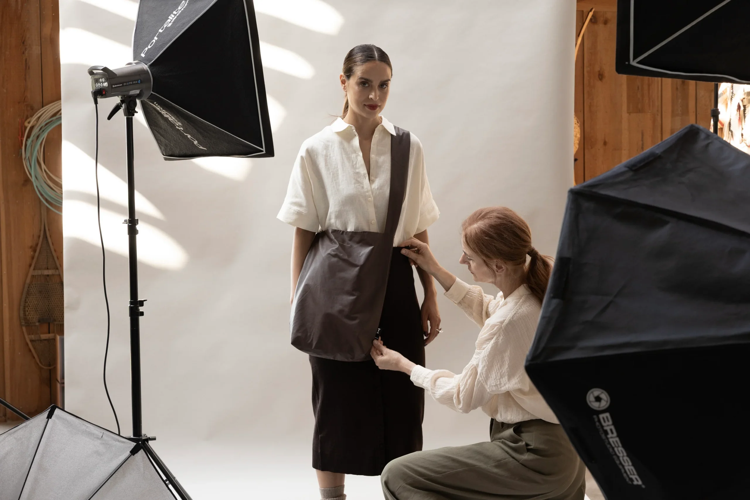 A woman poses in front of a white backdrop holding a Feel Good Bag - Chicory Coffee, dressed in a two-tone outfit, while another woman adjusts her clothing. Studio lights and equipment are visible around them in the photo studio. Gate21.ee