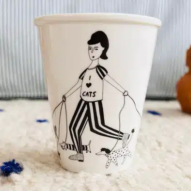 The I love Cats Cup is a white mug with a black and white illustration of a person in an "I love cats" shirt walking three cats on leads. Great for Tallinn shopping or as a unique gift shop find. Features a light background with blurred stripes. Gate21.ee