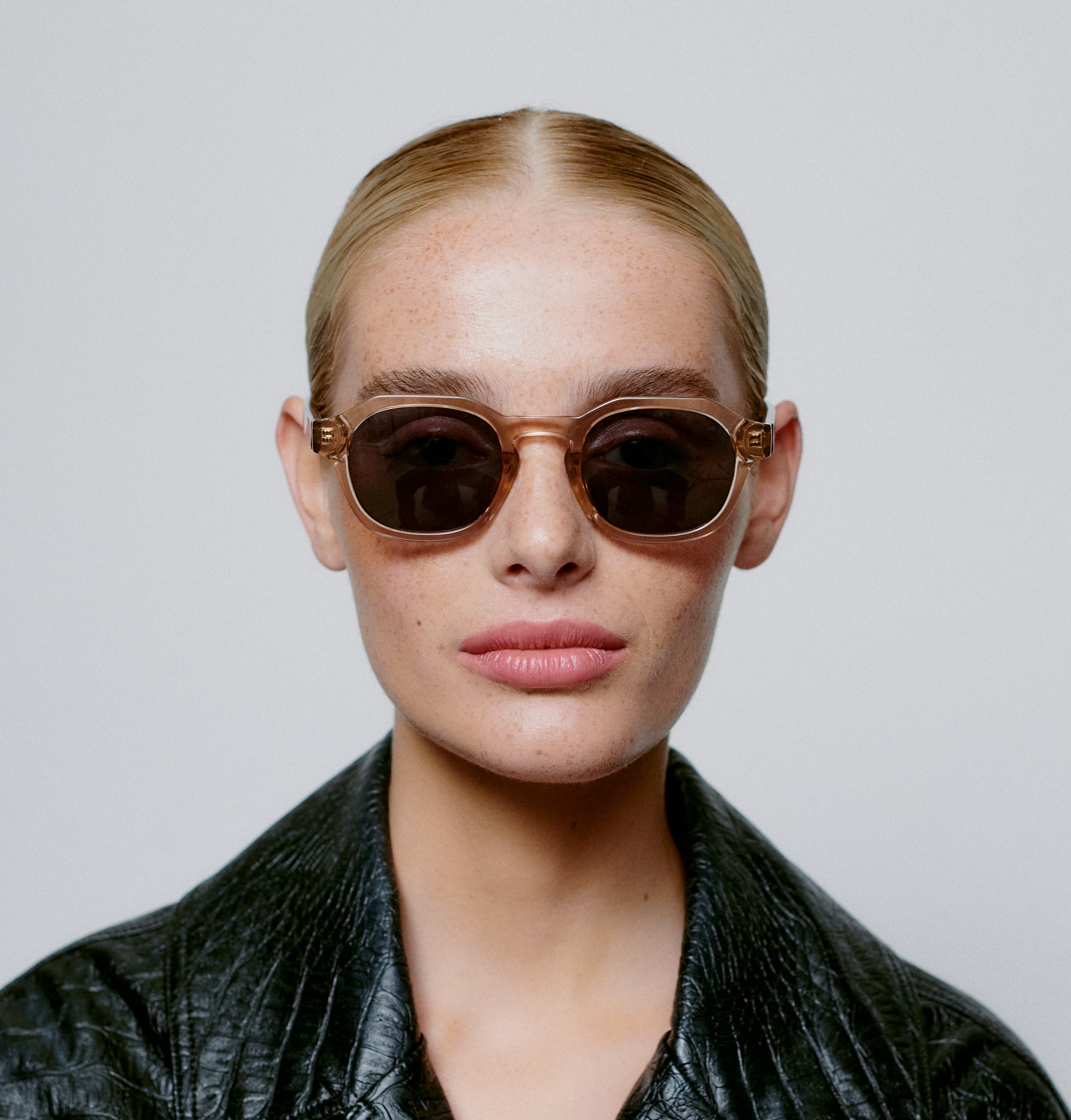 A person with slicked-back blonde hair, round sunglasses, and a textured black jacket faces the camera against a light background—embodying the chic vibe of ZAN - Champagne. Gate21.ee
