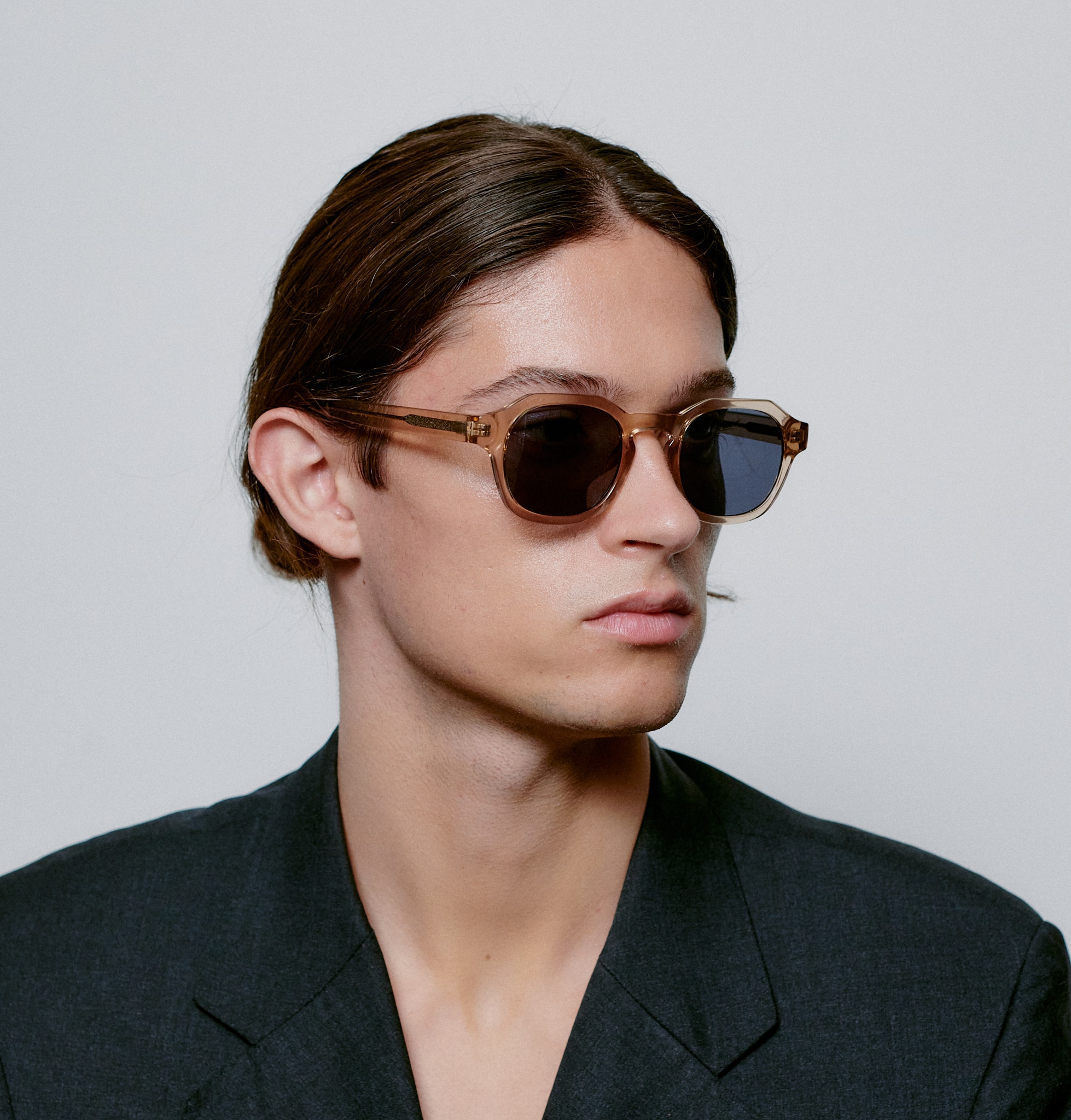 A person with long brown hair pulled back, wearing brown sunglasses and a dark blazer, poses against a plain background—perfect inspiration for unique ideas featuring the ZAN - Champagne from a Tallinn shopping concept shop. Gate21.ee