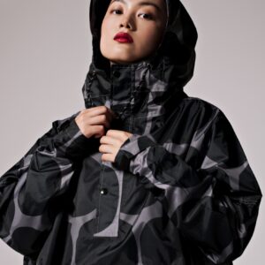 Wearing the "Polka" black and grey hooded jacket with a bold abstract pattern inspired by Estonian design, a person with short hair and red lipstick looks at the camera. The neck is fastened; the background is plain and light. Gate21.ee
