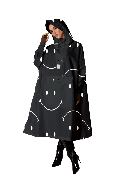 A woman models the Digi Spring black hooded rain poncho, featuring white smiley faces inspired by Estonian design, paired with black heeled boots against a plain white background. Gate21.ee