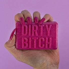 A hand with long, bright pink nails holds the DIRTY BITCH - Soap, a rectangular bar featuring Estonian design and bold embossing, set against a pink background—an original Tallinn shopping find. Gate21.ee