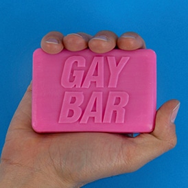 A hand displays the GAY BAR - Soap, a rectangular pink bar embossed with its name, set against a blue background—a playful piece of Estonian design for your next Tallinn shopping trip. Gate21.ee