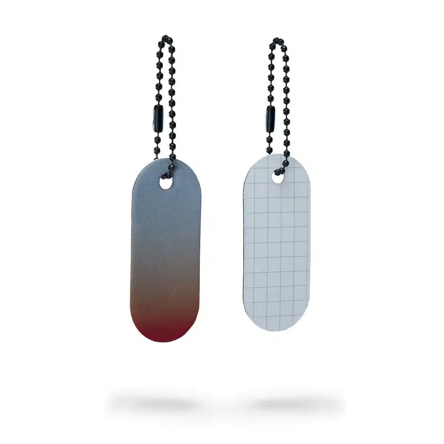 Two oval-shaped metal keyrings hang side by side on ball chains. The Reflective Tag – Red Fade boasts a grey surface with a red edge, standing out against a crisp white background—perfect for gift shops or concept stores. Gate21.ee