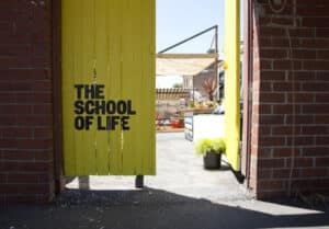 A bright yellow wooden gate with THE SCHOOL OF LIFE opens between two red brick walls, revealing a curated outdoor area with seating, potted plants, and the Compatibility Game displayed in a concept shop structure. Gate21.ee