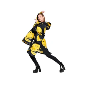 A person models the Smiley rain poncho from RAINKISS, featuring bold yellow smiley faces, paired with black leggings and platform boots against a white background. Gate21.ee