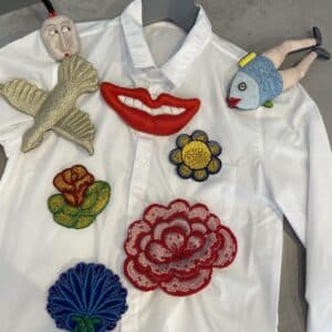 The Riivo Brooch - Large is displayed on a grey surface with vibrant appliqu&eacute;s&mdash;lips, flowers, a fish, bird, and human face&mdash;creating an artful scene from a curated concept store. Gate21.ee