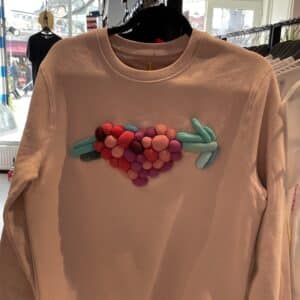 The Riivo Brooch - Large, a textured, multicoloured heart design of fabric spheres with an arrow through it, hangs in an Estonian concept store with clothing rails and a window visible in the background. Gate21.ee