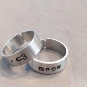 Two Kedrina aluminium rings &ndash; K&Otilde;IK rest on a grey surface. One shows a small bone symbol, the other is stamped with "meow." Find these playful pieces at our concept store, showcasing unique Estonian design for gift shops. Gate21.ee