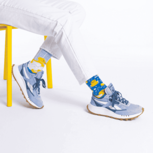 Seated on a yellow chair, someone sports Fisher's Tale blue socks featuring yellow rubber duck patterns with white trousers and light blue trainers—the perfect quirky find for your go-to concept store or gift shop. The background is white. Gate21.ee